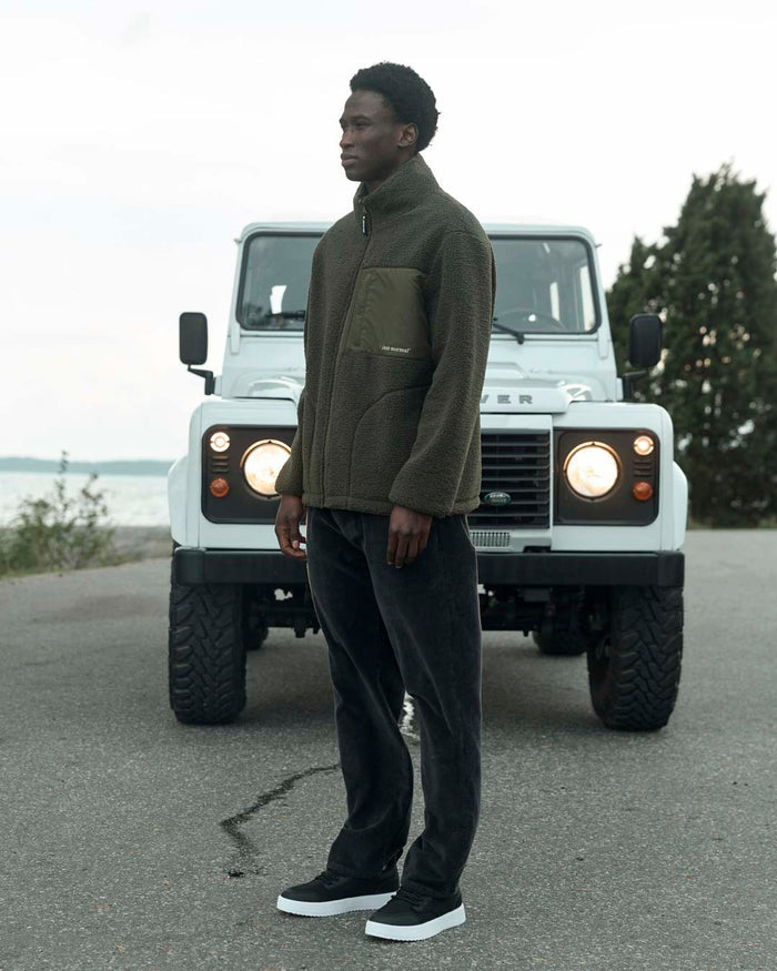A man standing in front of a white off-road vehicle, wearing The Reversible Pile Jacket in forest green and dark pants. The image highlights the pile side of the jacket, featuring a chest pocket with the c'est normal logo.