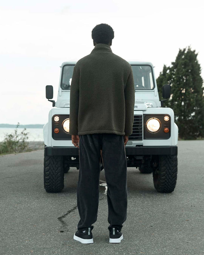 A back view of a man wearing The Reversible Pile Jacket in forest green by c'est normal, standing outdoors in front of a white off-road vehicle with its headlights on.