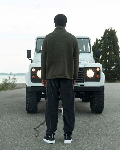 A back view of a man wearing The Reversible Pile Jacket in forest green by c'est normal, standing outdoors in front of a white off-road vehicle with its headlights on.