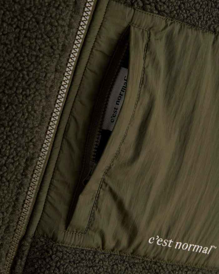 A detailed close-up of The Reversible Pile Jacket in forest green, focusing on the main zipper and the zippered chest pocket. The image showcases the contrast between the textured pile material and the smooth pocket fabric featuring the white c'est normal logo.