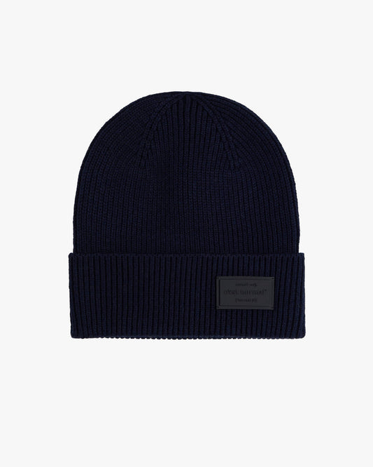 The Ribbed Beanie in navy blue, laid flat on a light grey background. The image shows the ribbed knit texture and the tone-on-tone rubber tag with the embossed c'est normal logo on the folded brim.