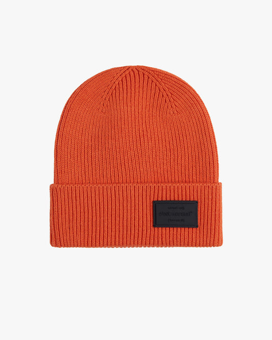 The Ribbed Beanie in orange, laid flat on a white background, highlighting its ribbed knit texture and the embossed c'est normal logo on a black rubber tag on the brim.