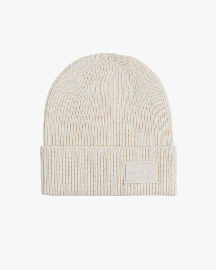 The Ribbed Beanie in cream, laid flat against a white background. The image features a clear view of the ribbed knit and the c'est normal logo embossed on a tone-on-tone rubber tag on the brim.