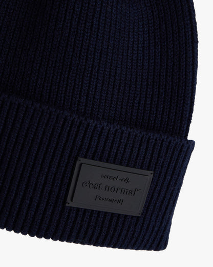 A detailed close-up of The Ribbed Beanie in navy, focusing on the tone-on-tone rubber tag on the brim with the embossed c'est normal logo. The image also showcases the classic ribbed knit material.