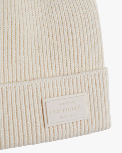 A detailed close-up of The Ribbed Beanie in cream, focusing on the ribbed knit texture and the tone-on-tone rubber tag with the embossed c'est normal logo on the brim.