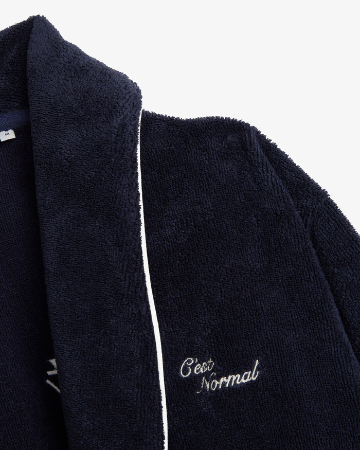 A detailed close-up of The Robe in navy, focusing on the soft cotton blend fabric and white piping. The image also showcases the embroidered c'est normal logo on the chest.