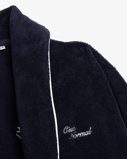 A detailed close-up of The Robe in navy, focusing on the soft cotton blend fabric and white piping. The image also showcases the embroidered c'est normal logo on the chest.