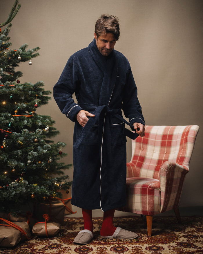 A man tying the belt of The Robe in navy with white piping, standing next to a decorated Christmas tree and a plaid armchair. The image highlights the cozy, oversized fit and the embroidered c'est normal logo on the chest.