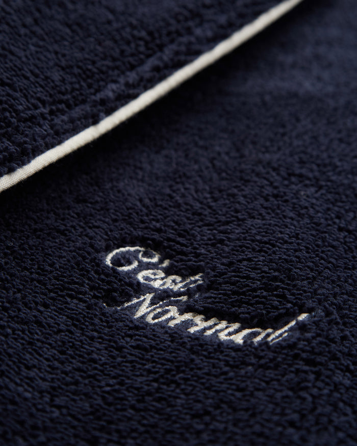 A detailed close-up of The Robe in navy, focusing on the white embroidered c'est normal logo and white piping. The image highlights the soft, textured cotton blend material.