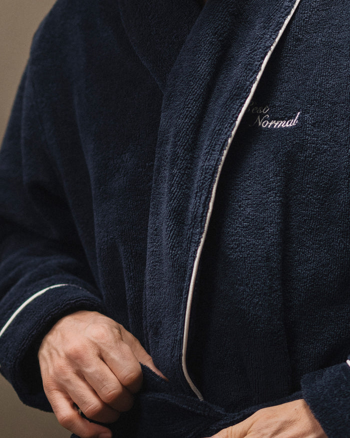 A close-up of a person wearing The Robe in navy, highlighting the soft fabric, white piping, and the embroidered c'est normal logo on the chest.