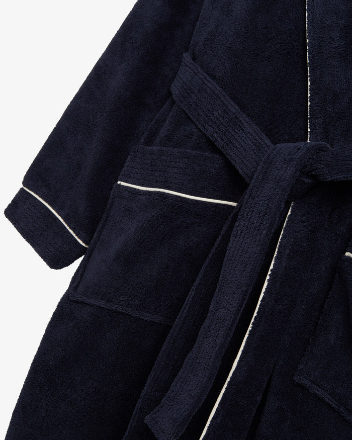 A close-up shot of The Robe in navy, laid flat against a white background. The image highlights the soft cotton-poly blend fabric, the tie belt, the large front pocket, and the classic white piping detail.