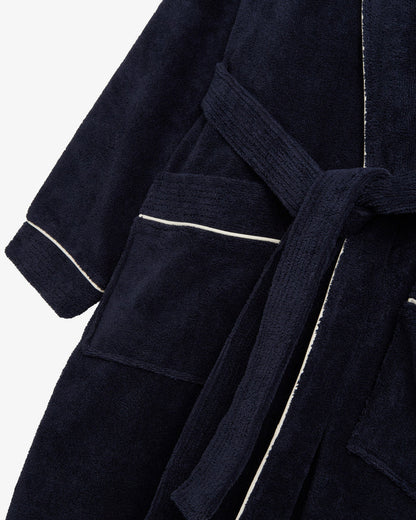 A close-up shot of The Robe in navy, laid flat against a white background. The image highlights the soft cotton-poly blend fabric, the tie belt, the large front pocket, and the classic white piping detail.