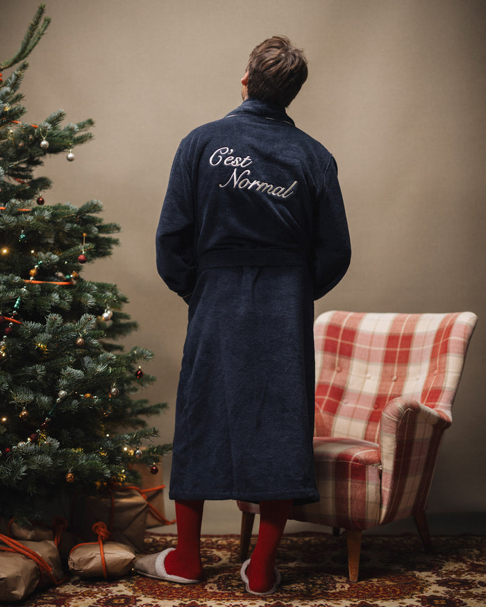 A back view of a man wearing The Robe in navy, standing next to a decorated Christmas tree and a plaid armchair. The image highlights the white embroidered c'est normal logo on the back of the robe.