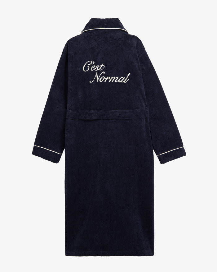 A back view of The Robe in navy, laid flat on a white background. The image highlights the white piping on the collar and cuffs, the matching belt, and the large embroidered c'est normal logo on the back.
