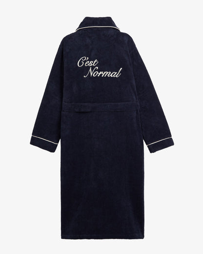 A back view of The Robe in navy, laid flat on a white background. The image highlights the white piping on the collar and cuffs, the matching belt, and the large embroidered c'est normal logo on the back.