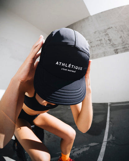 A person adjusting The Running Cap in black, looking down to highlight the front of the cap. The cap features the text 'ATHLÉTIQUE' and the c'est normal logo in white.