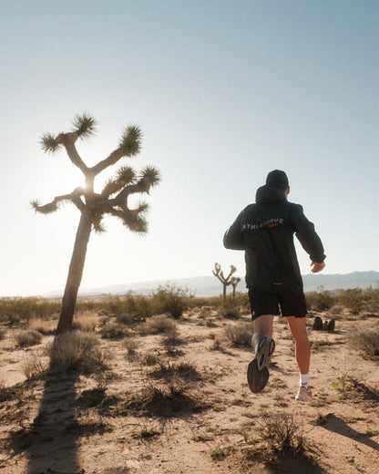 A man running in a desert landscape with Joshua trees, viewed from behind. He is wearing The Running Jacket in black, which features "ATHLÉTIQUE" and the c'est normal logo on the back.