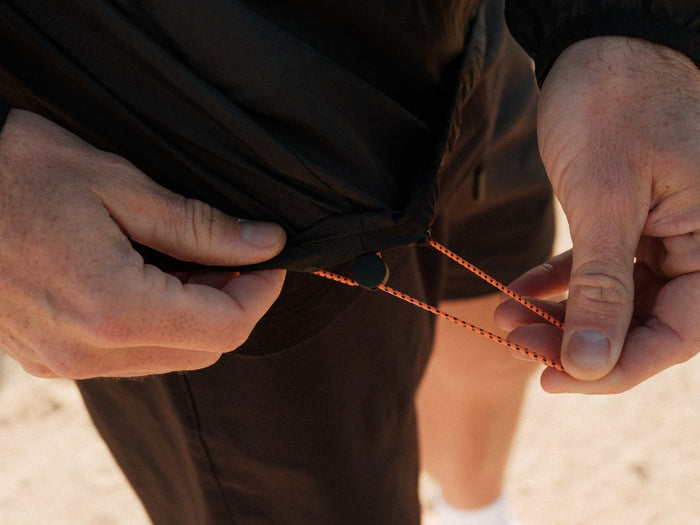A close-up shot of a person adjusting the orange and black drawstring on the hem of The Running Jacket in black.