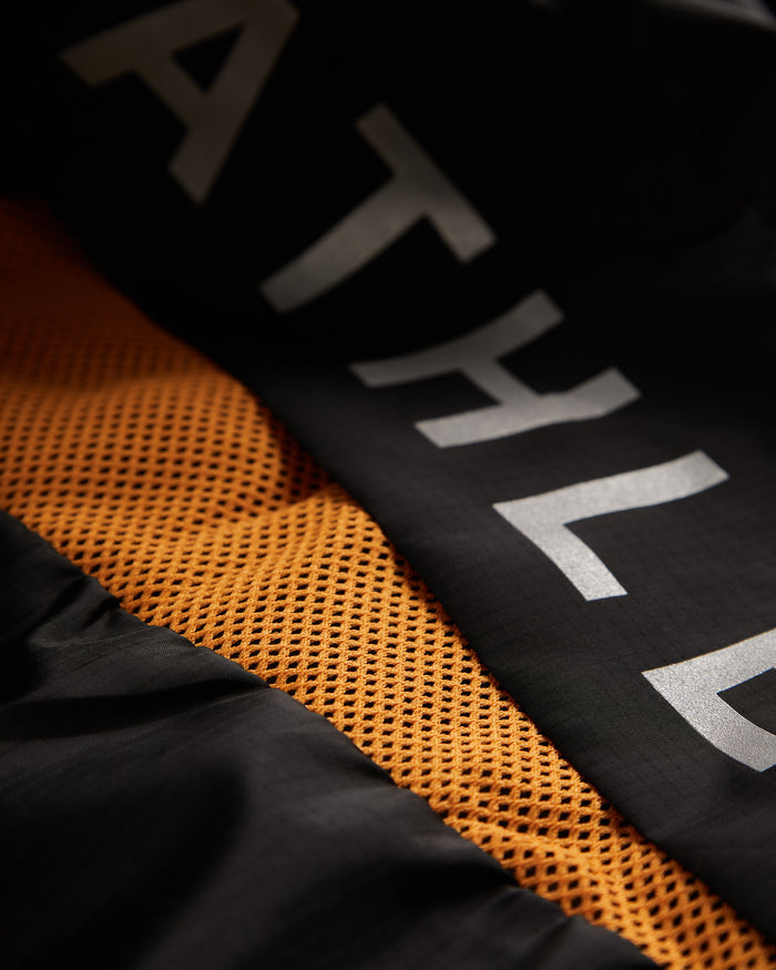 A close-up shot of The Running Jacket in black, highlighting the reflective ATHLÉTIQUE logo on the back, the orange mesh ventilation panel, and the black fabric.