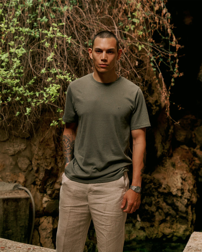 The Sanremo Linen Tee in mulled green — worn with beige trousers against a rocky wall