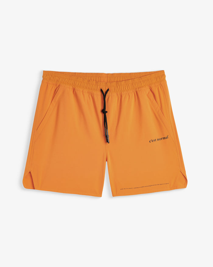 A front view of The Shorts in orange peel, laid flat on a white background. The image highlights the elasticated waistband with a black drawstring, side pockets, notched hems, and the printed c'est normal logo.