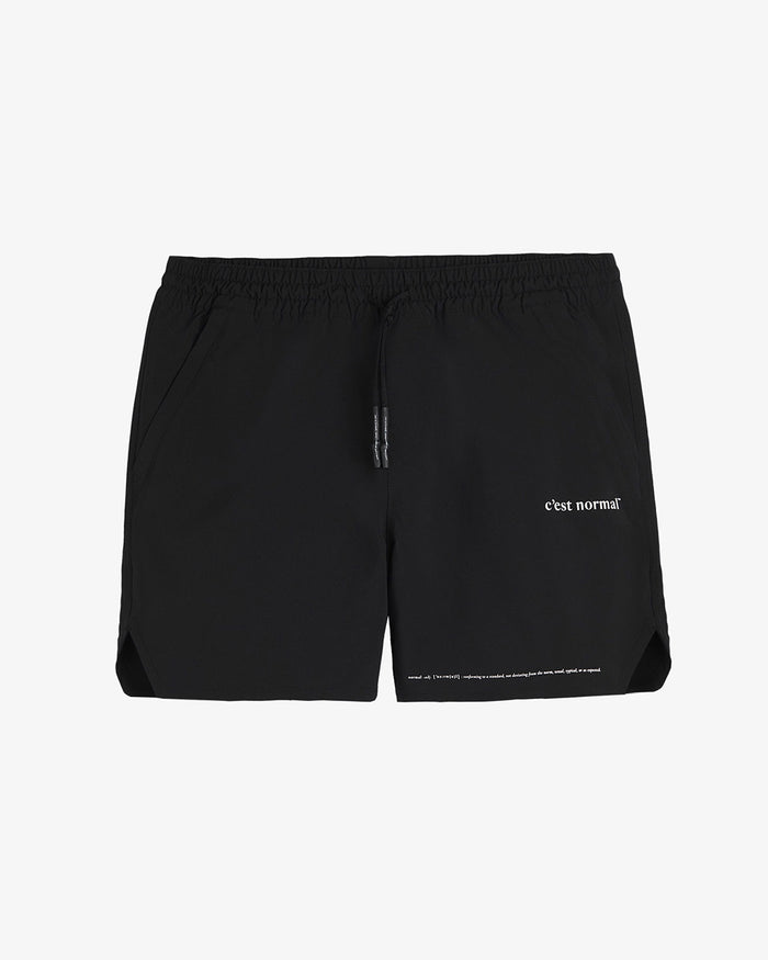 A front view of The Shorts in black, laid flat on a white background. The image highlights the elastic waistband with drawstrings, zippered side pockets, notched hem, and the white c'est normal logo on the left leg.