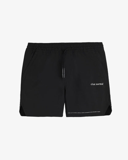 A front view of The Shorts in black, laid flat on a white background. The image highlights the elastic waistband with drawstrings, zippered side pockets, notched hem, and the white c'est normal logo on the left leg.