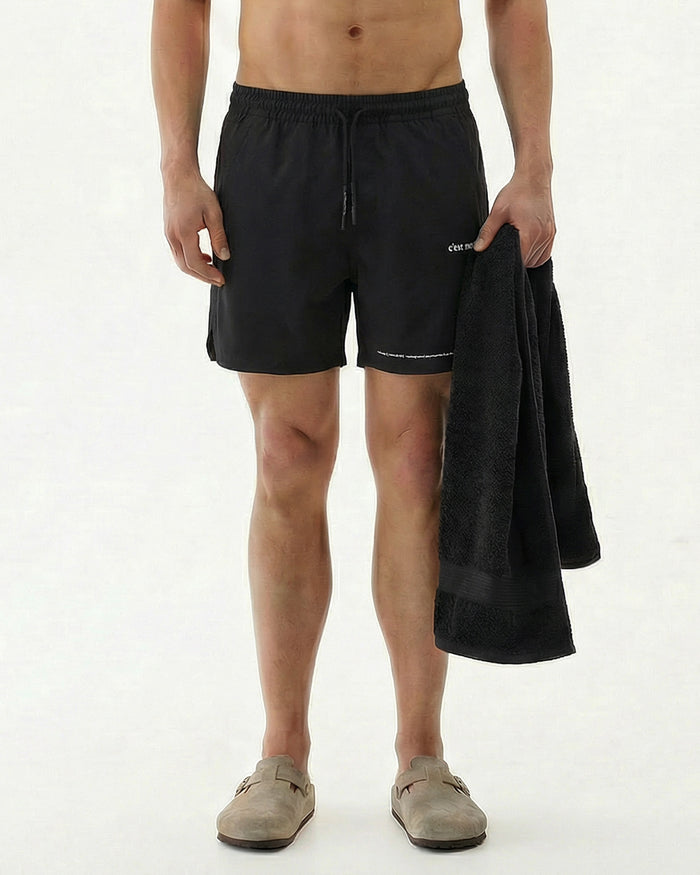 A model standing on a white background wearing the black The Shorts, holding a black towel. The image highlights the c'est normal logo on the left leg.