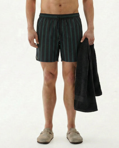 A shirtless model standing against a white background, wearing The Shorts in Phantom Stripe with a dark green and black striped pattern. He holds a black towel in his left hand and wears beige slip-on shoes.