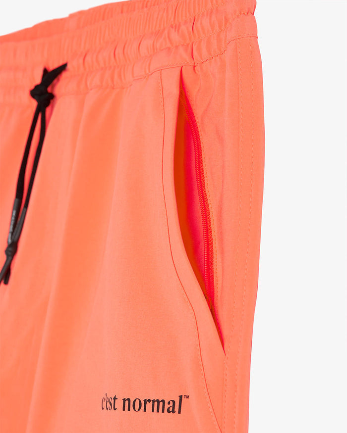 A close-up view of The Shorts in orange, showing the zippered side pocket, black drawstring with aglets, and the black c'est normal logo near the hem.