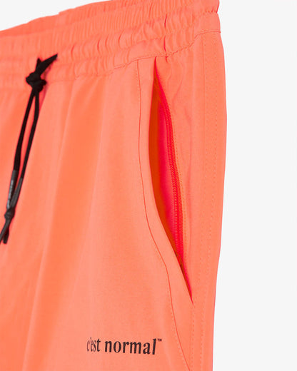 A close-up view of The Shorts in orange, showing the zippered side pocket, black drawstring with aglets, and the black c'est normal logo near the hem.
