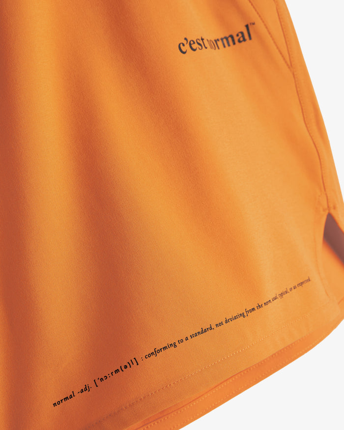 A close-up shot of The Shorts in orange peel, highlighting the smooth, stretch fabric and the side slit on the hem. The image features the printed c'est normal logo near the top pocket and a dictionary definition of 'normal' printed along the bottom hem.