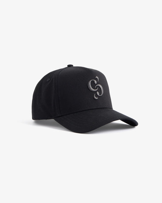 A front angled view of The Snapback Cap in black, resting on a white background. The image highlights the dark embroidered c'est normal mirror logo on the front panel.