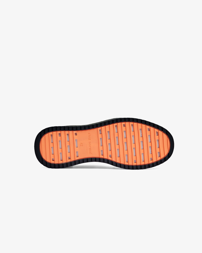 A bottom view of The Sneakers 2.0 in black, laid flat on a white background. The image highlights the original orange EVA rubber grid sole featuring the c'est normal logo.