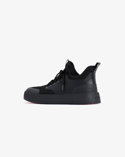 A side profile view of The Sneakers 2.0 in black on a white background. The image highlights the mesh upper, rubber accents, thick midsole, and the embossed c'est normal logo on the heel.
