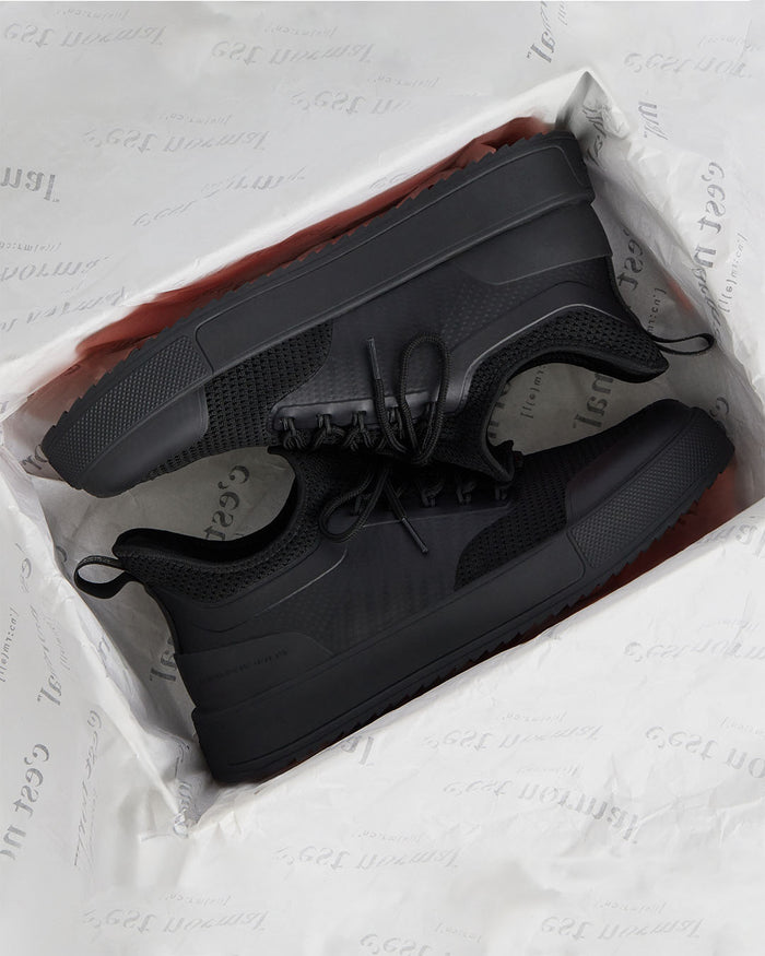 A pair of The Sneakers 2.0 in black, resting inside a shoe box lined with white tissue paper featuring the c'est normal logo. The image highlights the tone on tone mesh upper, custom lace loops, and thick running midsole.