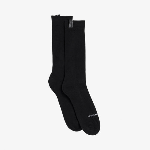 A pair of The Sock in black, laid flat on a white background. The image highlights the ribbed mid-calf length, the cuff tag, and the white c'est normal logo across the toe.