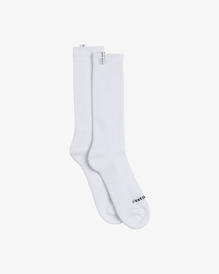A pair of The Sock in white, laid flat on a white background. The image highlights the mid-calf ribbed design and the c'est normal logo on the cuff tag and across the toe.