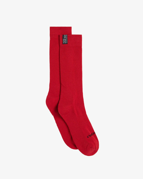 A pair of The Sock in red, laid flat on a white background. The image highlights the ribbed mid-calf design, the black cuff tag with the c'est normal logo, and the c'est normal logo across the toe.