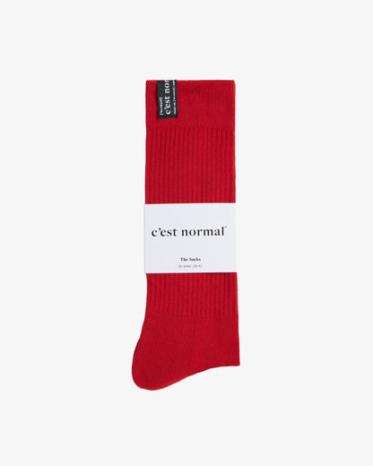 A pair of The Sock in red, laid flat on a white background, wrapped in a white paper sleeve. The image highlights the ribbed texture and the black cuff tag featuring the c'est normal logo.