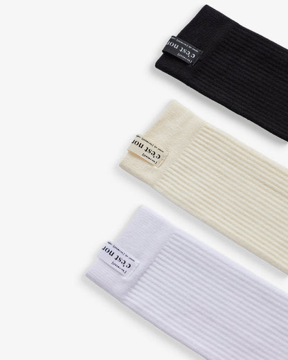 A top-down view of three The Sock variations in black, cream, and white, laid flat diagonally on a white background. The image highlights the ribbed texture and the cuff tags featuring the c'est normal logo.
