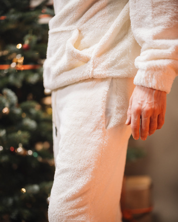 A close-up shot of a person wearing The Teddy Pant and matching hoodie in a creamy winter white, highlighting the soft, fleecy texture and the side pocket. A Christmas tree is visible in the background.