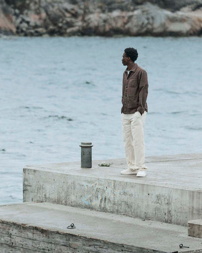A man standing on a concrete pier looking out over the water, wearing The Workwear Overshirt in brown with white pants.