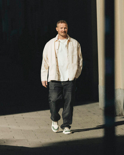 A man walking outdoors wearing The Workwear Overshirt in cream, styled open over a white t-shirt and dark jeans. The image highlights the relaxed fit and front patch pockets of the c'est normal overshirt.