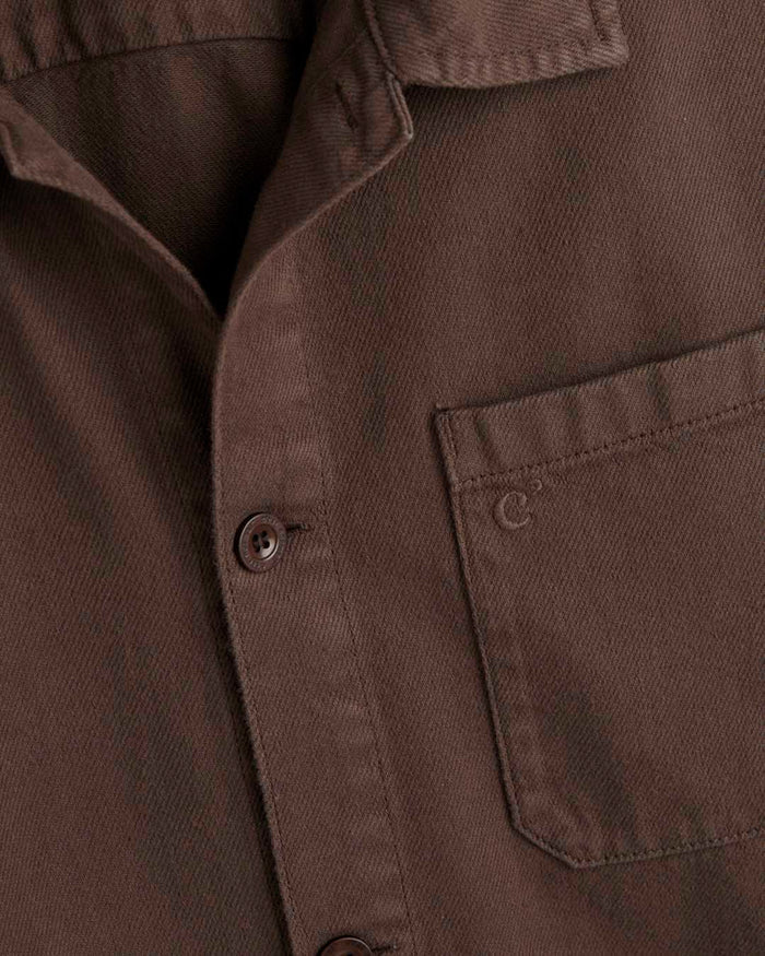 A detailed close-up of The Workwear Overshirt in brown, focusing on the left chest patch pocket with tone-on-tone c'est normal branding and the front button closure.