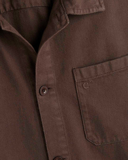 A detailed close-up of The Workwear Overshirt in brown, focusing on the left chest patch pocket with tone-on-tone c'est normal branding and the front button closure.