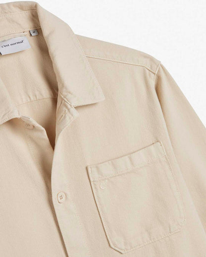 A close-up view of The Workwear Overshirt in cream, highlighting the collar, front buttons, and left chest patch pocket with subtle tone-on-tone branding. The inside neck label features the c'est normal logo.