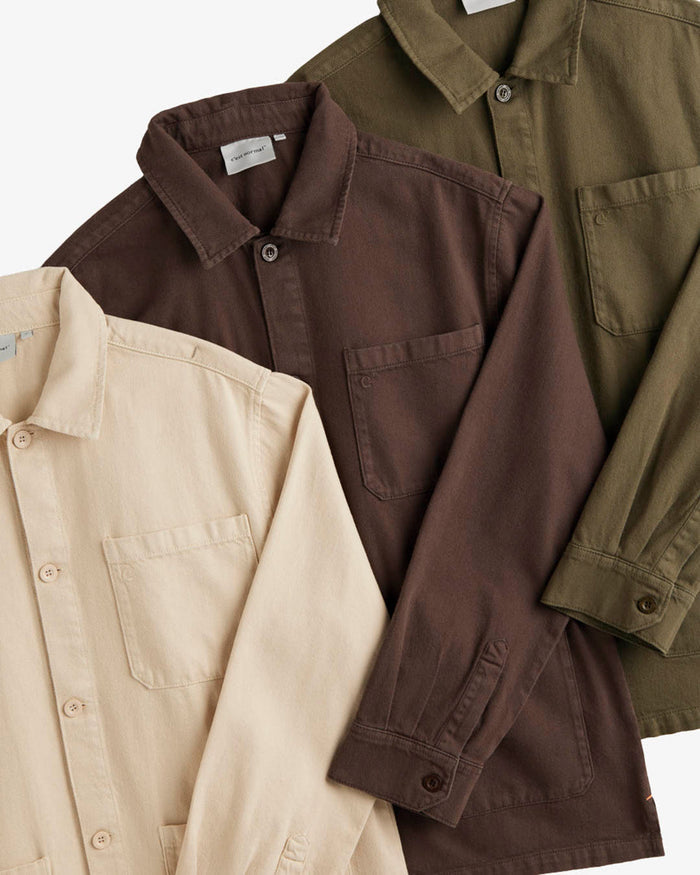 A close-up shot of three folded The Workwear Overshirts, laid flat in beige, brown, and olive green colors. The image highlights the collar, chest pockets with the c'est normal logo, and button details.