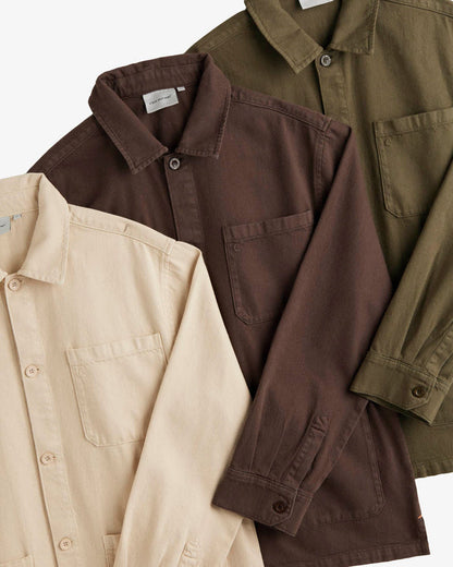 A close-up shot of three folded The Workwear Overshirts, laid flat in beige, brown, and olive green colors. The image highlights the collar, chest pockets with the c'est normal logo, and button details.