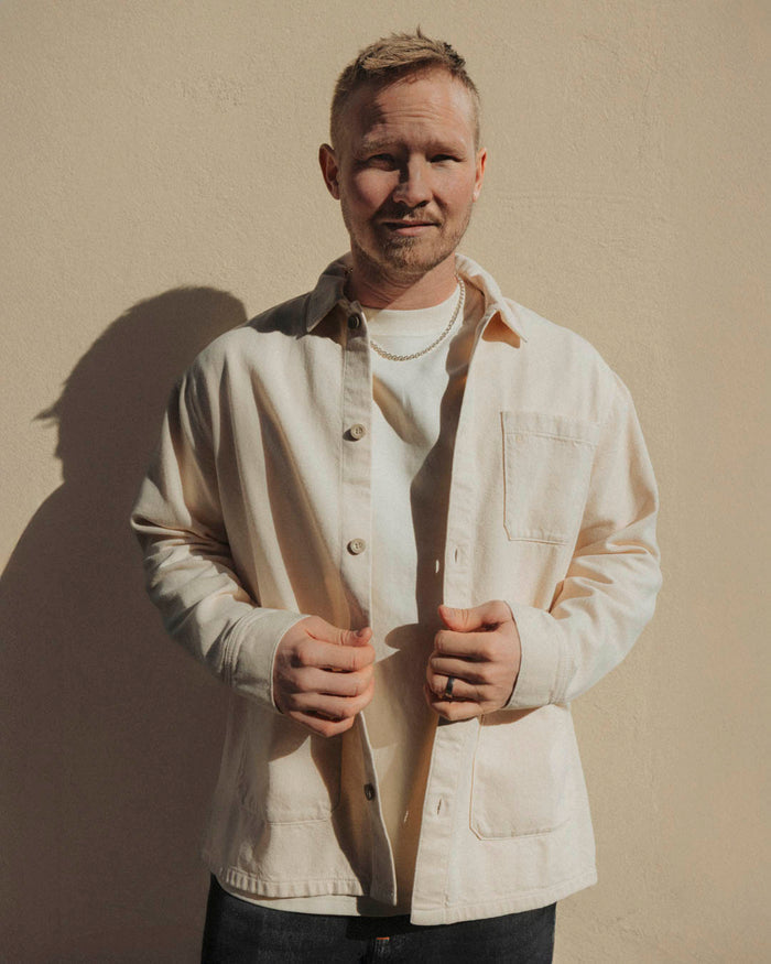 A man wearing The Workwear Overshirt in cream over a white t-shirt, standing outdoors in front of a beige wall. The image highlights the relaxed fit and patch pockets of the overshirt.